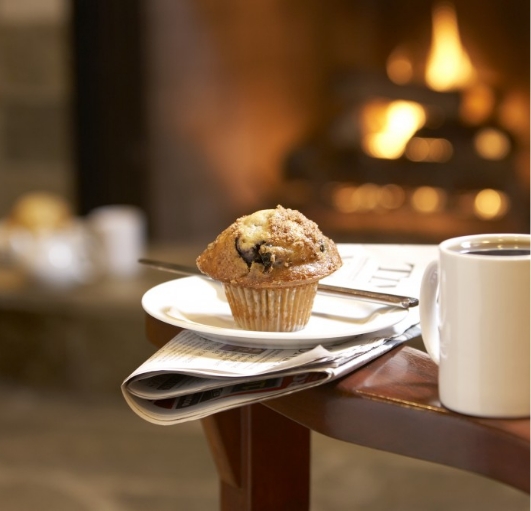 A muffin on a plate on top of a newspaper beside a full coffee cup on the arm of a chair in front of a fireplace.