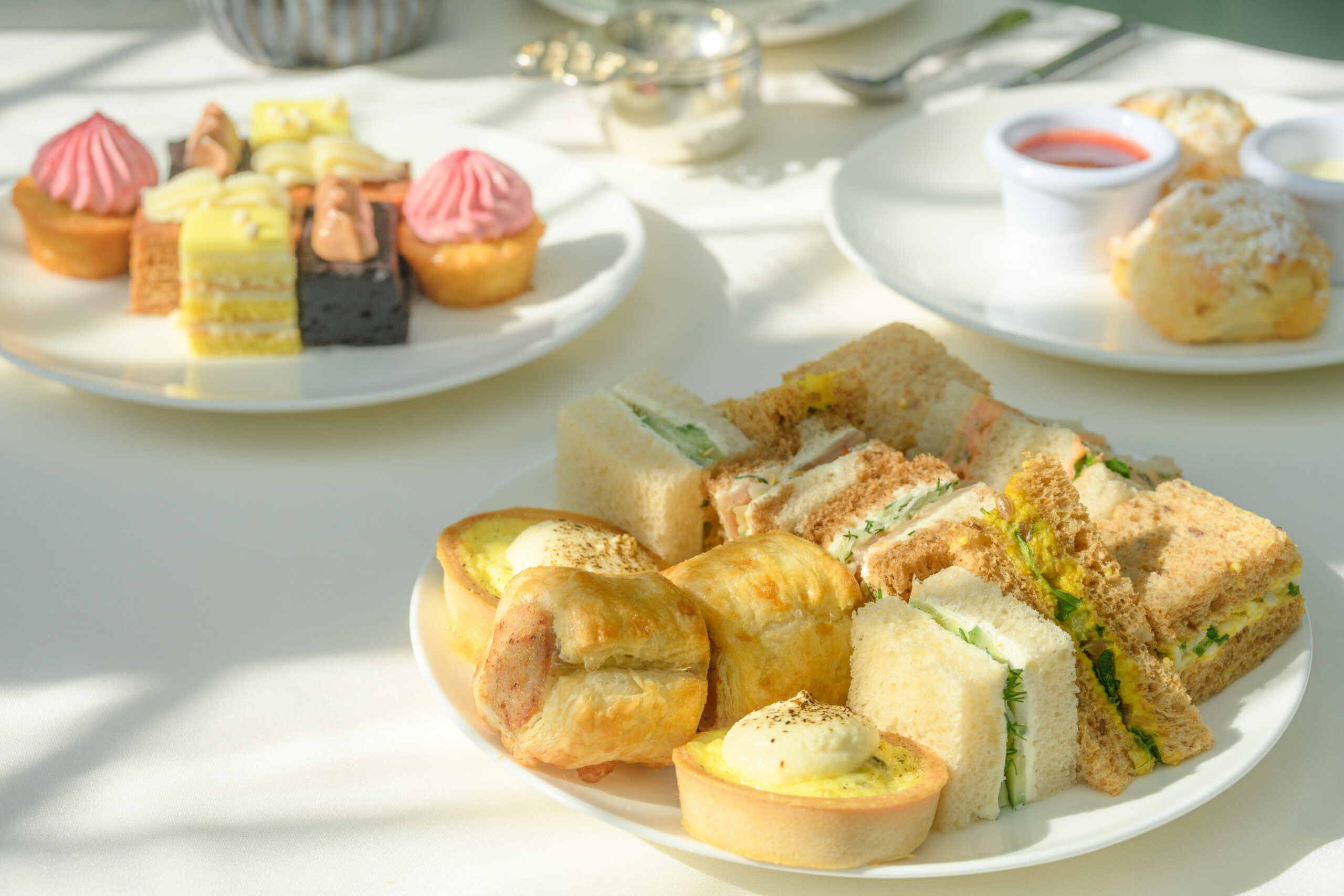 Tea sandwiches sit on a plate in the front of the frame. A plate filled with small desserts slightly blurred in the far left corner and a plate with two scones and jam sits blurred in the right hand corner.