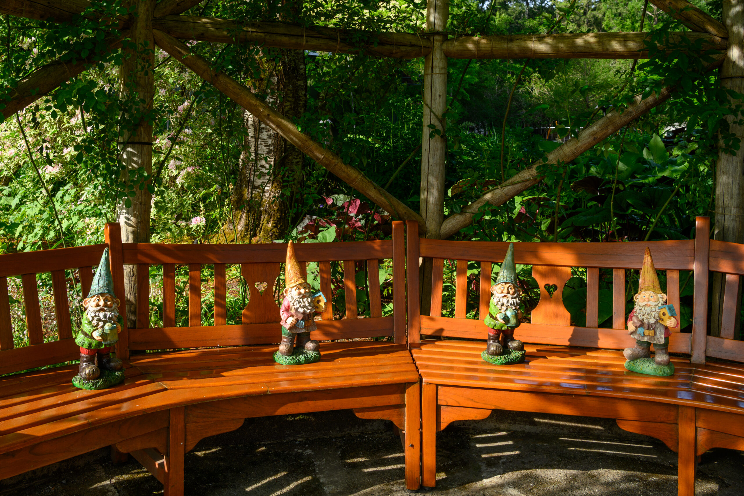 Three garden gnomes are placed on a wooden bench in a garden setting. Each gnome has a distinct design: one holds flowers, another has a fishing rod, and the third is depicted with a shovel. The bench is surrounded by greenery and wooden lattice structures
