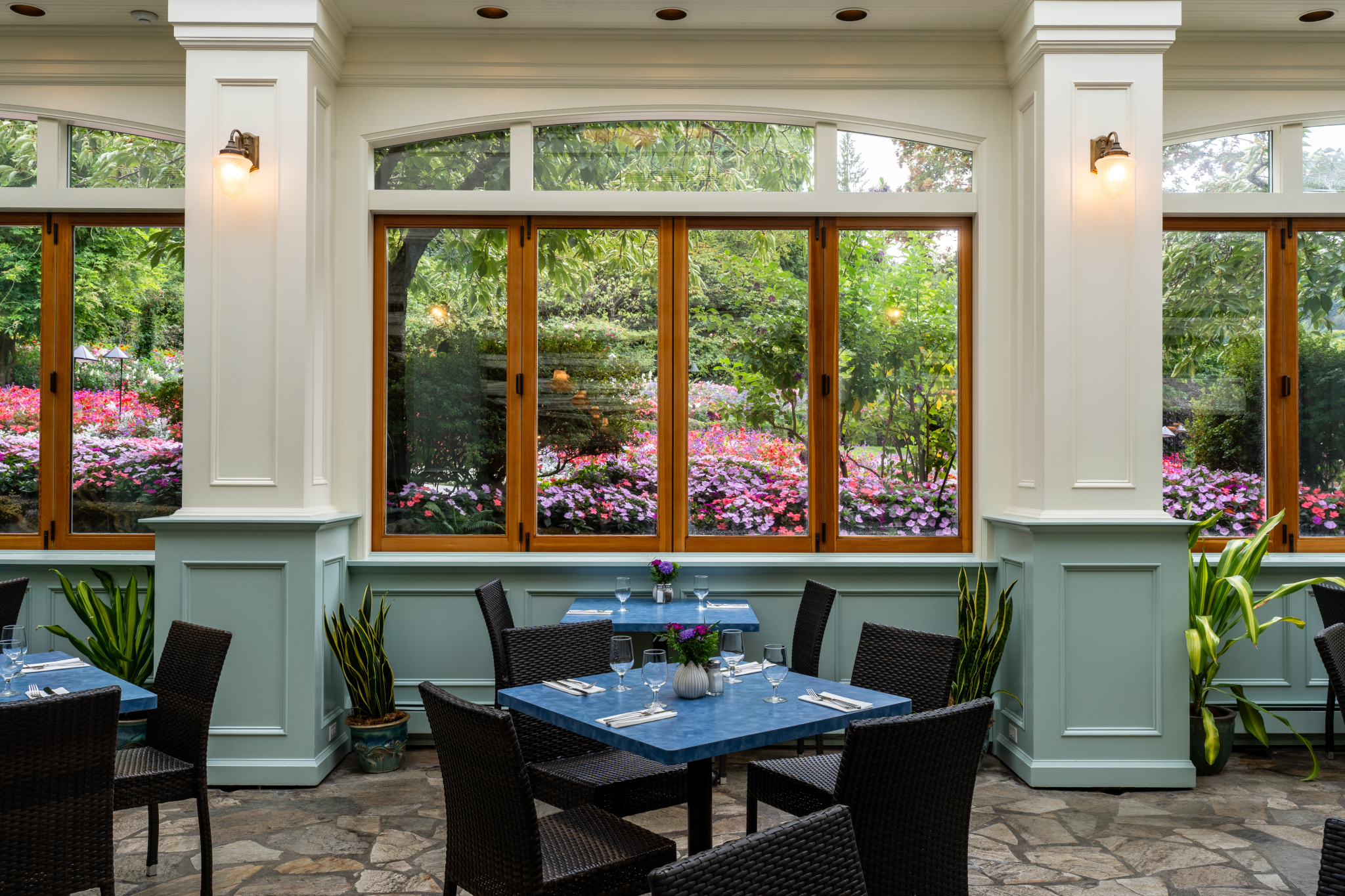 A bright dining area with large windows showcasing a vibrant garden filled with colorful flowers. The interior features light blue walls and stone flooring, with dark wicker tables and chairs arranged neatly. Potted plants are placed near the windows, adding greenery to the space
