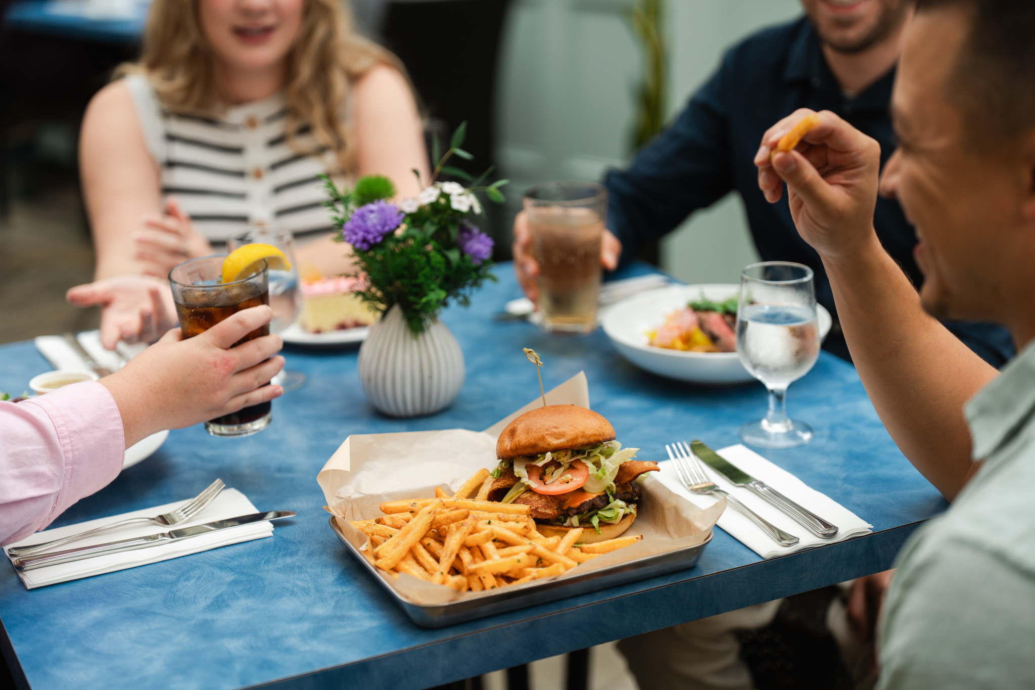 A group of four people is seated around a blue table in a restaurant. In the foreground, a plate holds a hamburger topped with lettuce, tomato, and a slice of onion, accompanied by a serving of golden French fries. A small vase with purple flowers is placed on the table, alongside glasses of water and soft drinks. The individuals are engaged in conversation, with one person raising a drink
