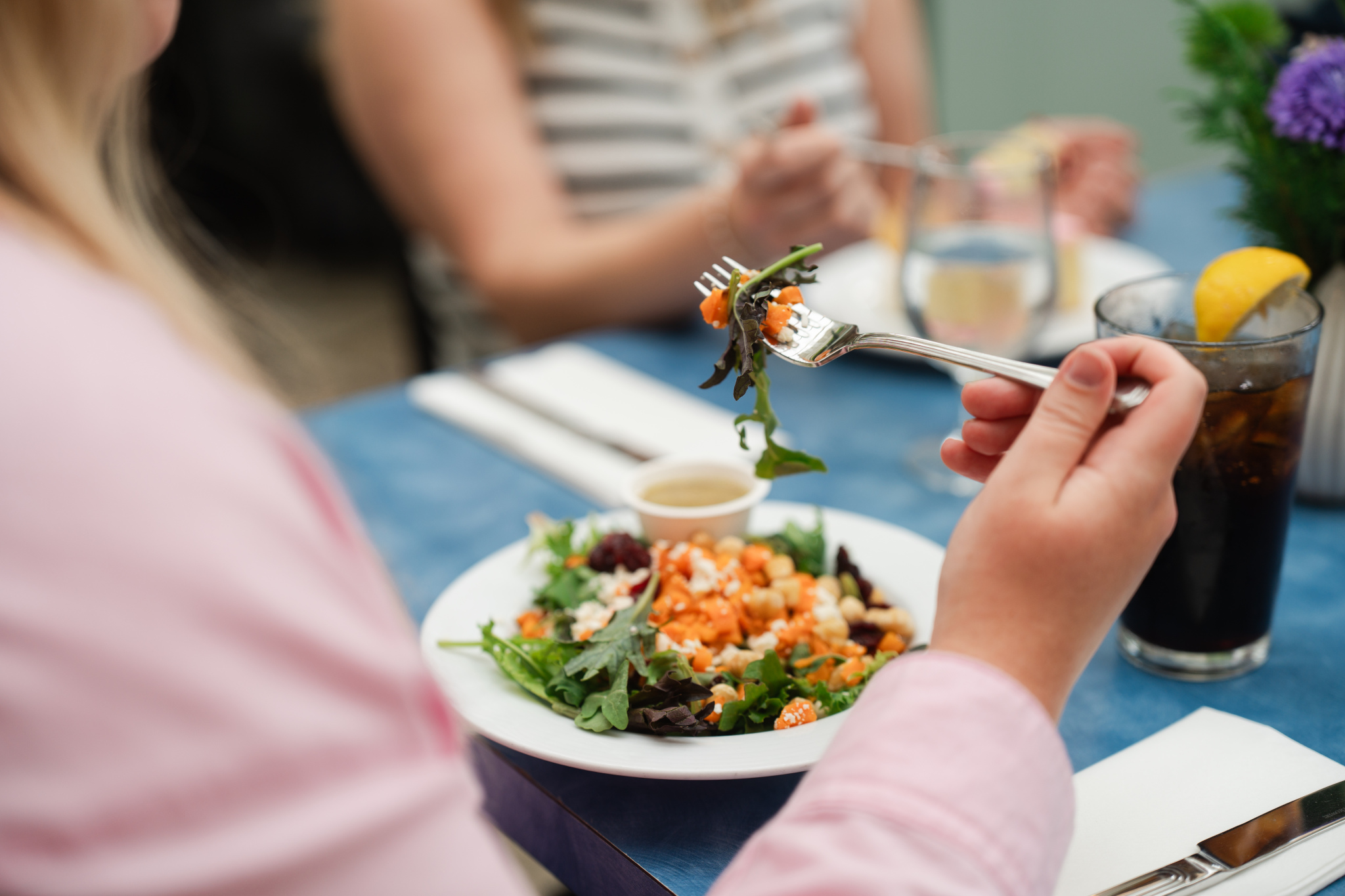 A person holding a fork with a piece of salad, featuring mixed greens, orange segments, and a light dressing. In the foreground, a plate of salad is visible, while a glass of dark beverage with a lemon slice is on the table. Another person is partially visible in the background