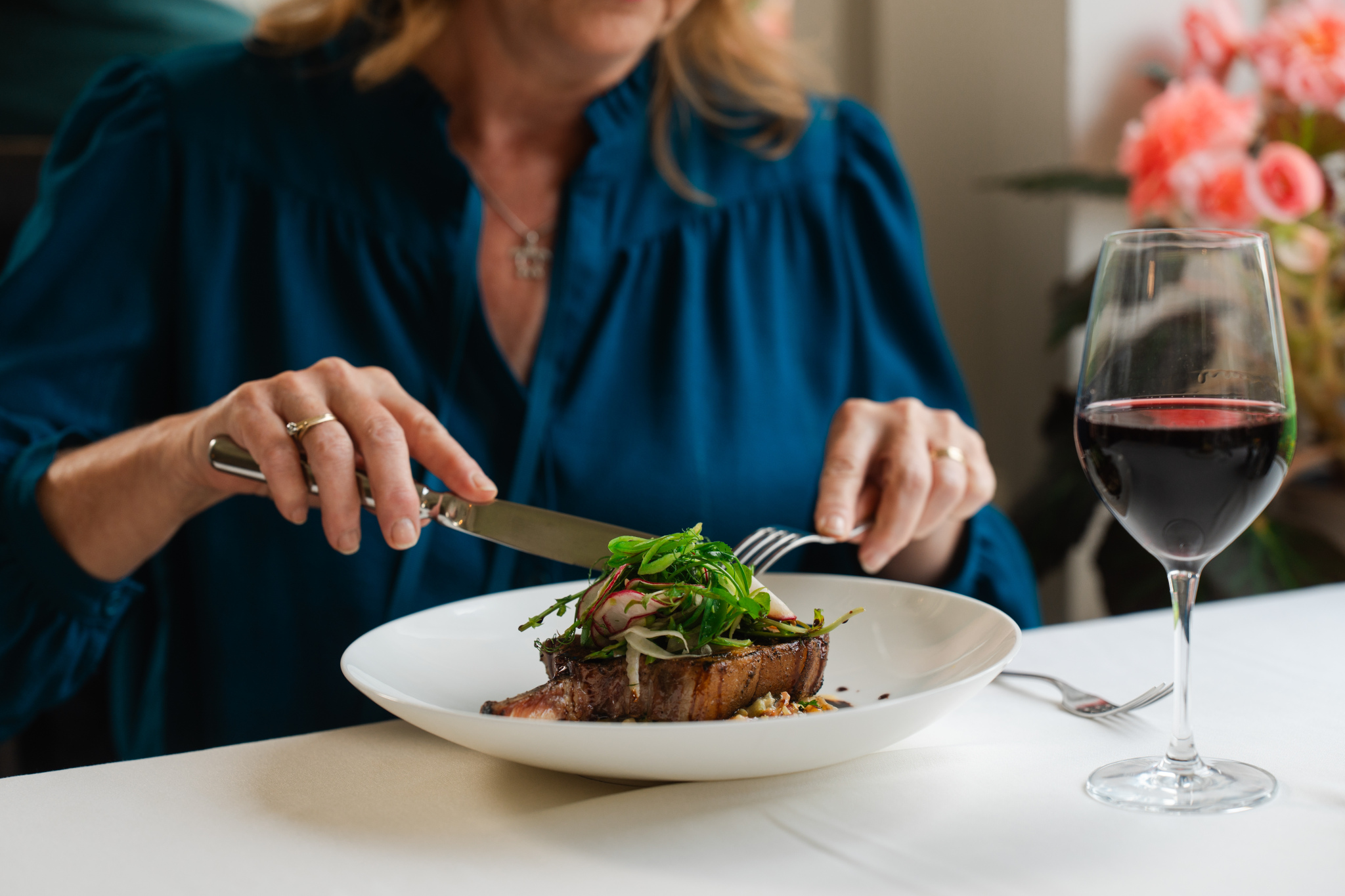 A woman in a blue blouse is cutting into a plate of steak topped with greens and garnishes. A glass of red wine is placed beside her on the table. The background features blurred pink flowers