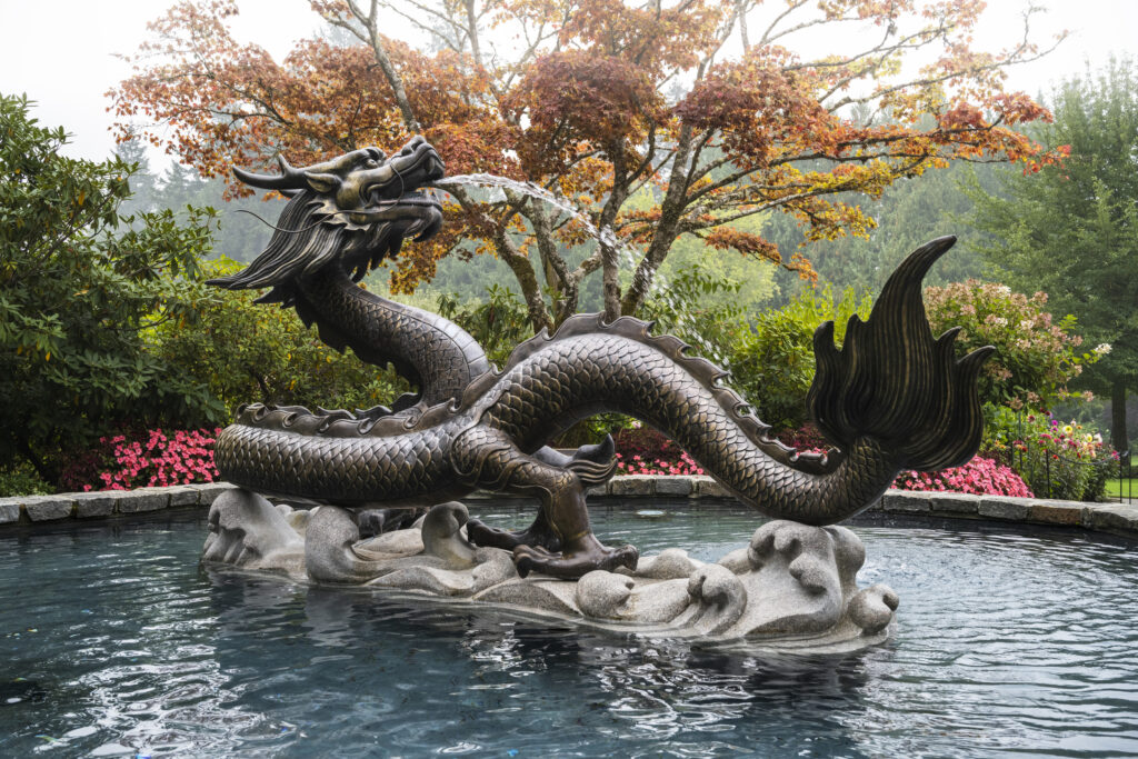 The Dragon Fountain on a hazy day with pink flowers and a leafy tree transitioning into autumn colours.