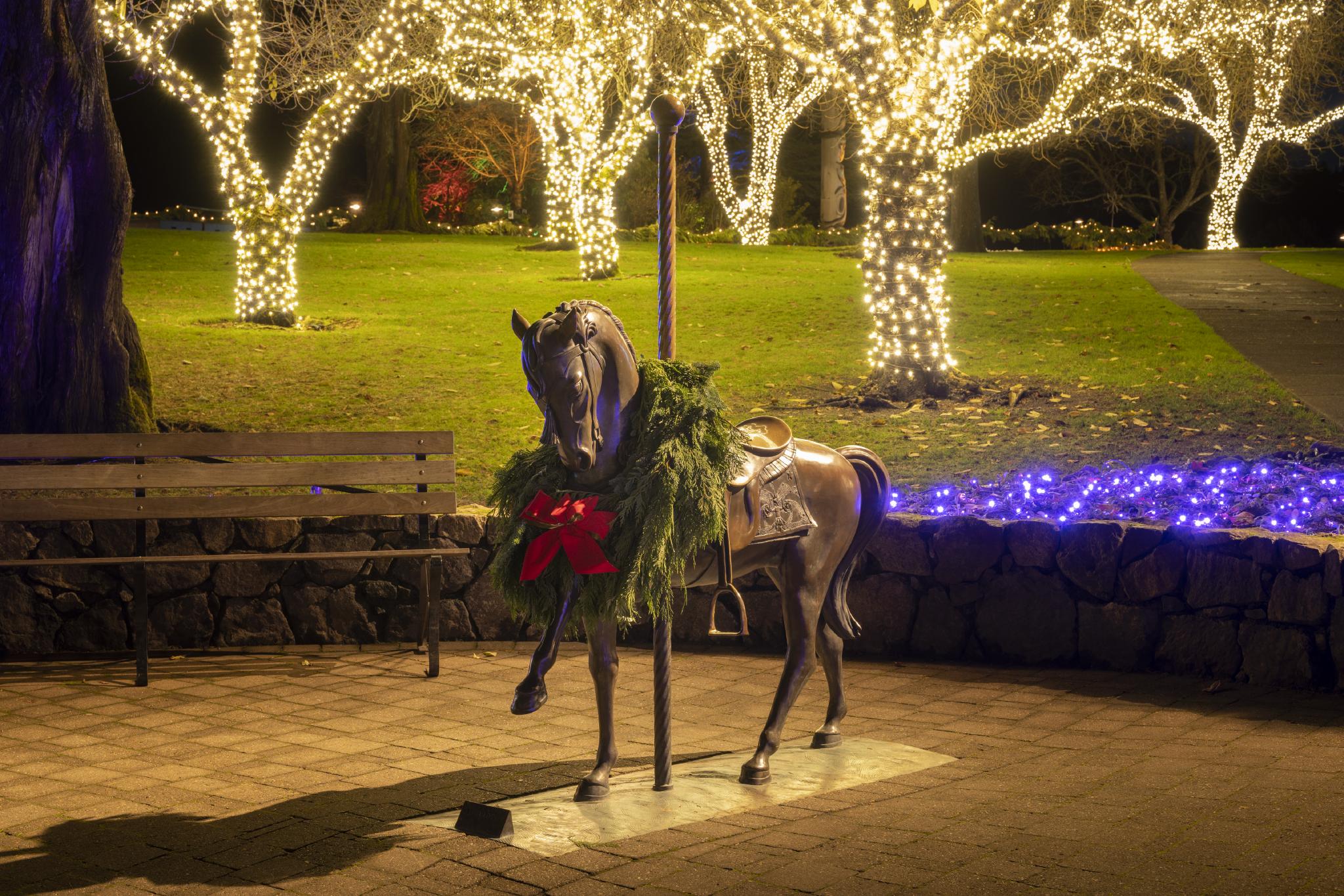 Statue en bronze d'un cheval de carrousel portant une couronne de Noël avec un nœud rouge avec, en arrière-plan, des arbres enveloppés de lumières de Noël.