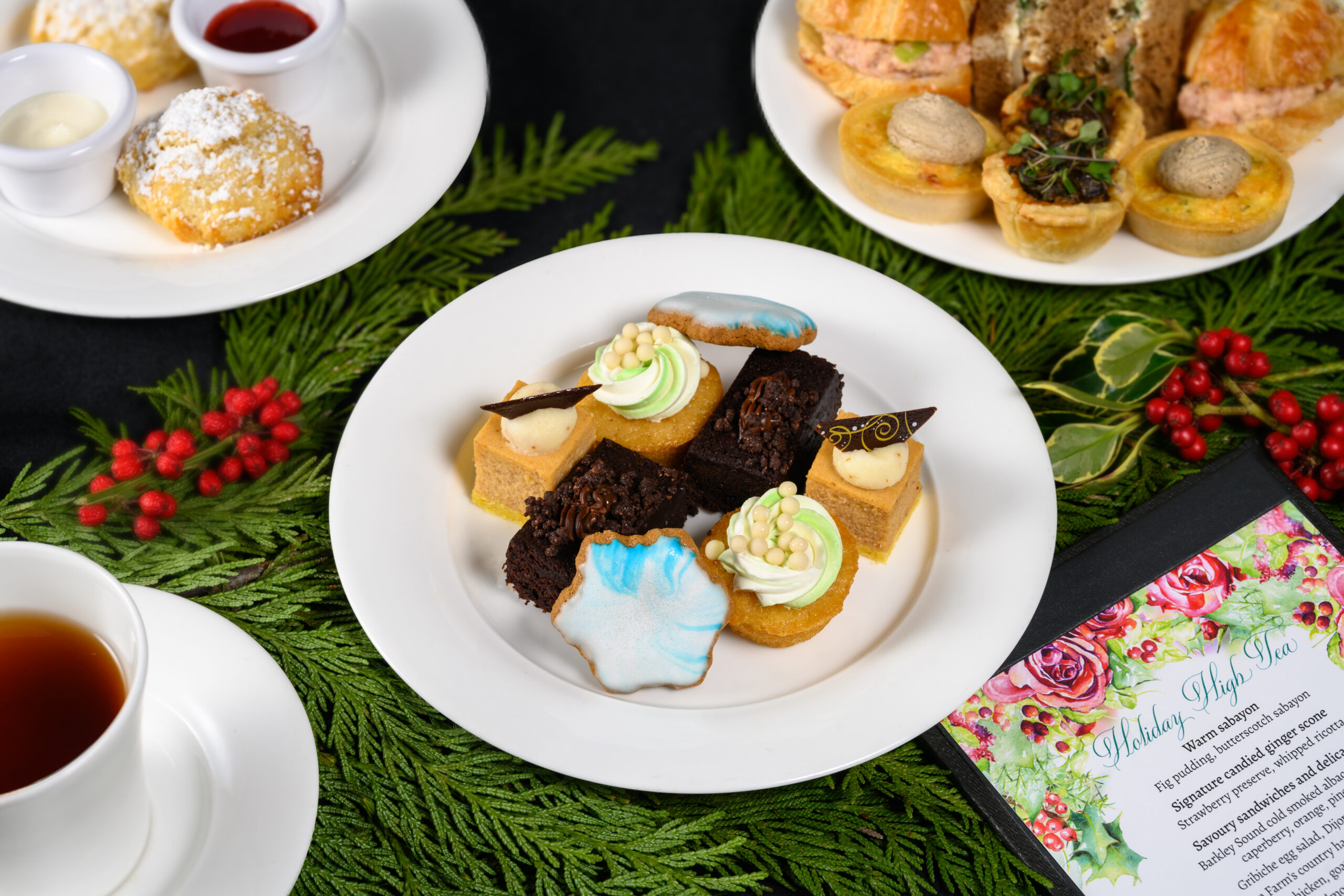 High Tea de Noël servi dans la salle à manger du restaurant. Trois assiettes blanches remplies de pâtisseries, de scones et de sandwichs au thé, avec une tasse à thé, une soucoupe et un menu. 