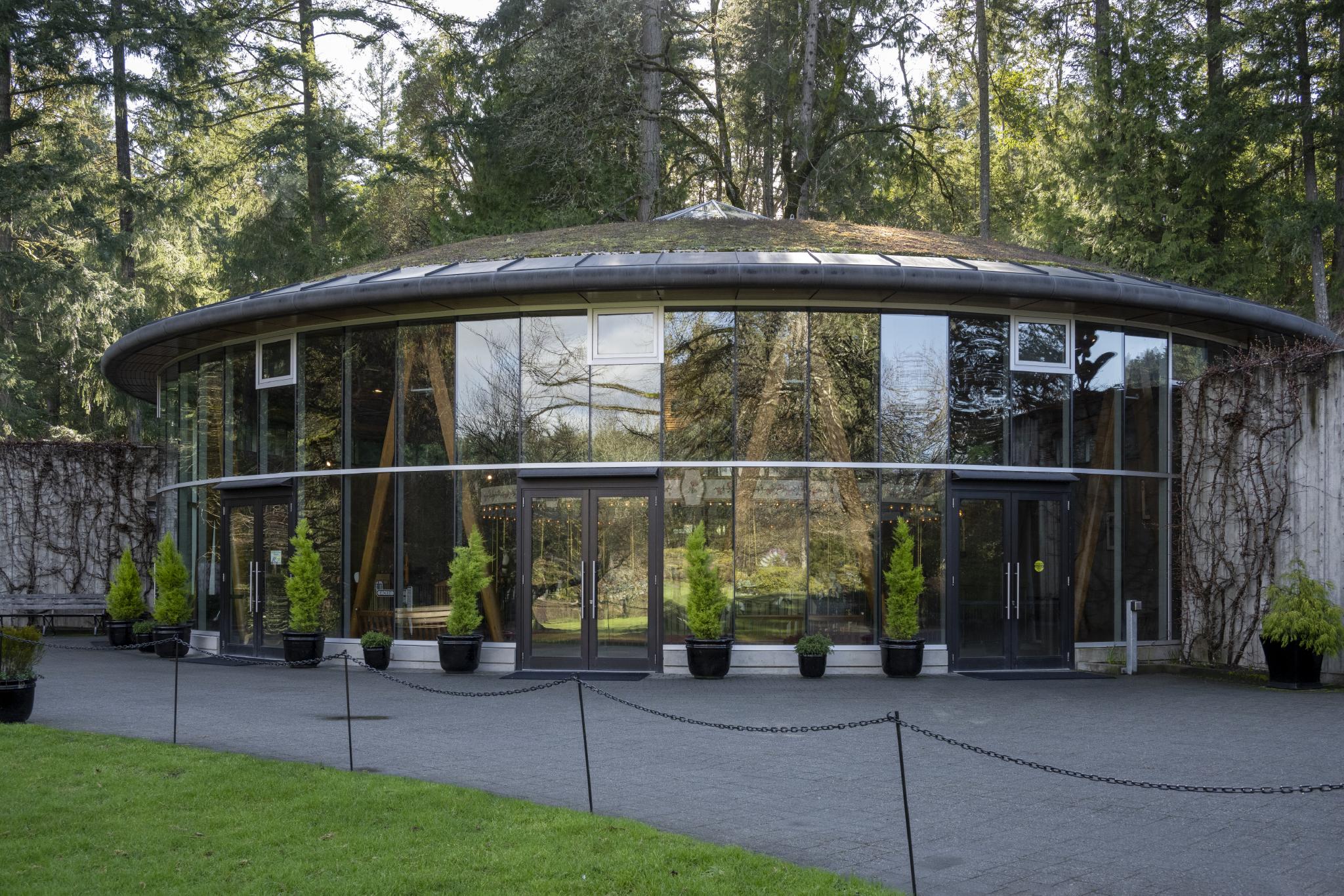 Large glass rounded building lined with potted hedges outside reflecting the surrounding landscape in the windows. The glow from a wooden carousel inside the building shines through the glass.