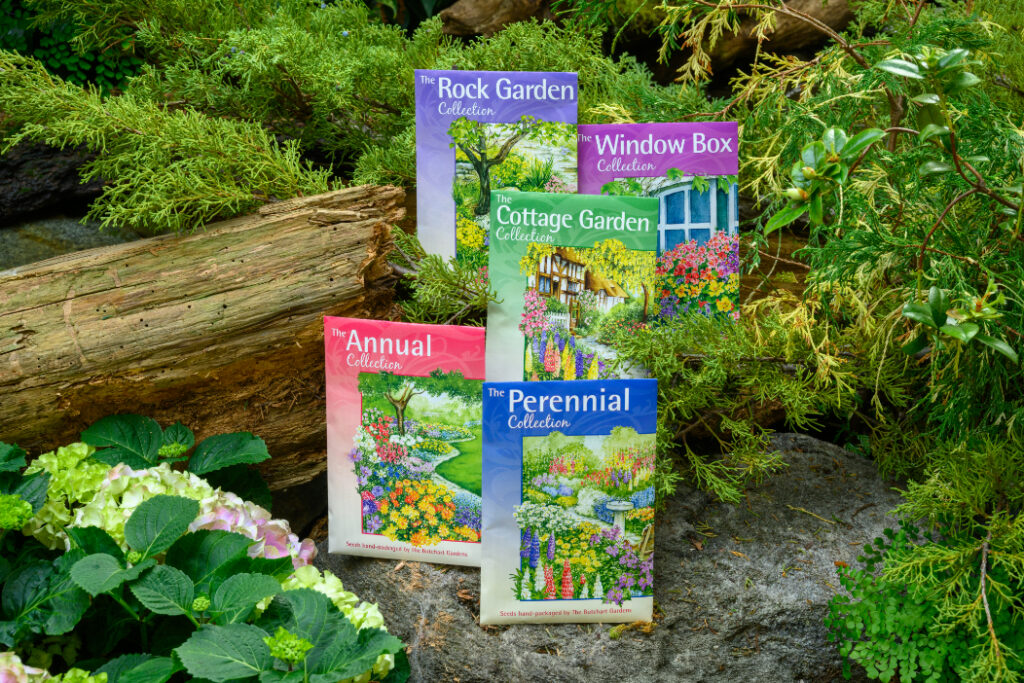 Five seed packages are displayed against a natural background of logs and greenery. The collections are "The Rock Garden," "Window Box," "Cottage Garden," "The Annual," and "The Perennial." Each cover features colourful illustrations of various garden scenes and plants