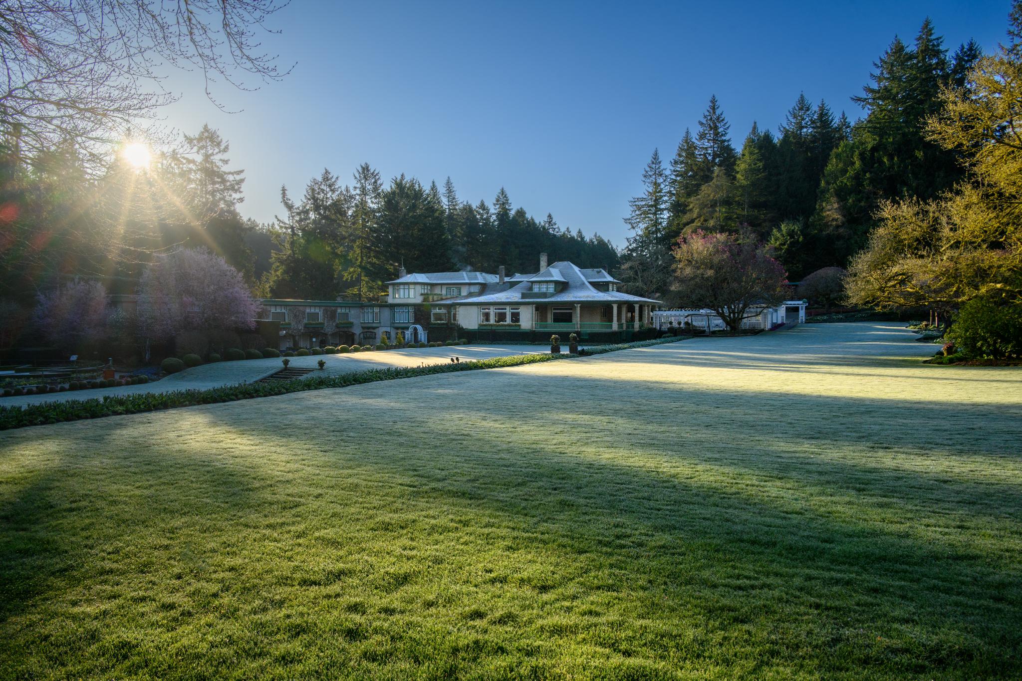 A large, modern house with a circular roof is set against a backdrop of evergreen trees. The lawn is covered in frost, with long shadows stretching across the grass. In the foreground, there are manicured flower beds and a pathway leading to the house. The scene is illuminated by soft morning sunlight