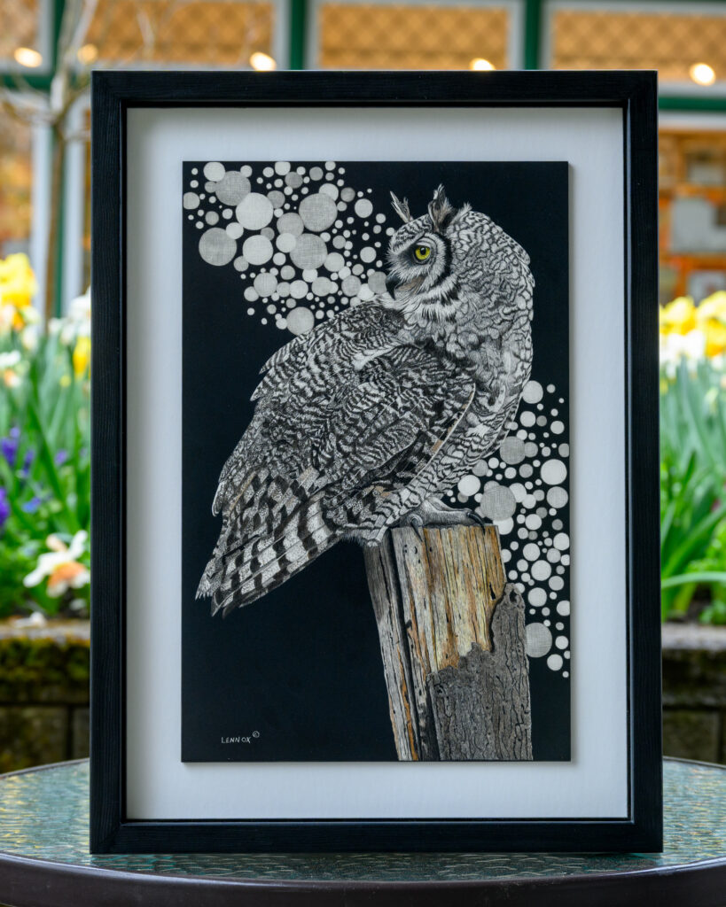A detailed illustration of a great horned owl (Bubo virginianus) perched on a wooden post. The owl is depicted with intricate feather patterns and a striking yellow eye. The background features a series of white and gray circular shapes, contrasting with the dark backdrop. The artwork is framed in a black frame and displayed on a table with blurred flowers in the background