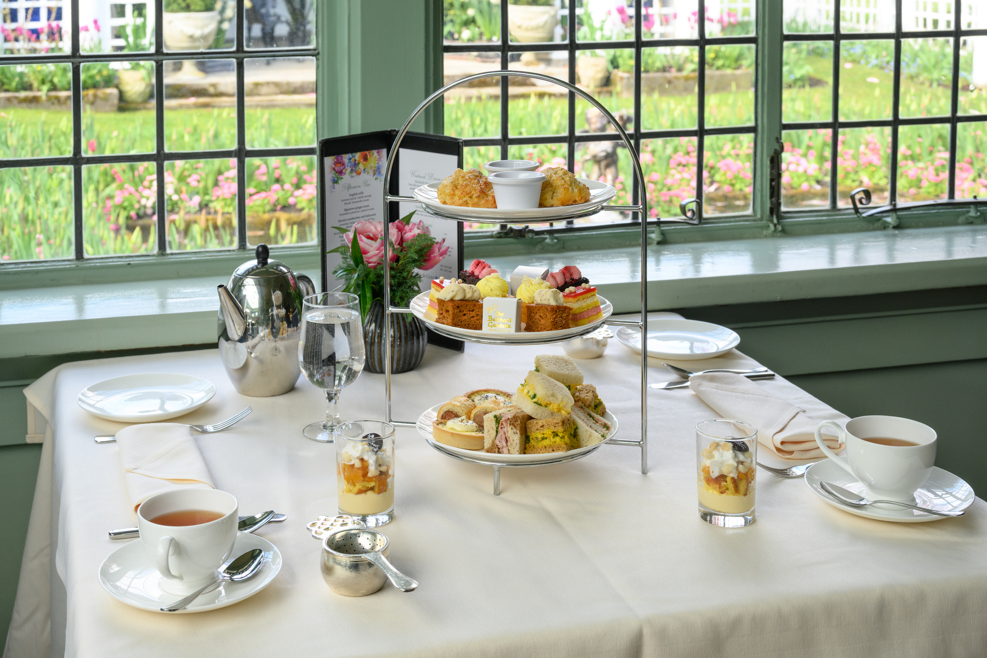 A tiered serving stand displays an assortment of pastries and sandwiches, including scones, fruit tarts, and small cakes. A teapot and teacups are set on a table covered with a cream-colored tablecloth. In the background, large windows reveal a garden with blooming flowers