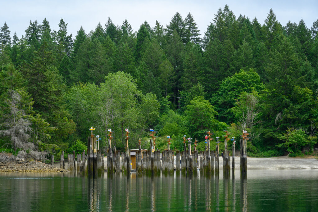 A series of weathered wooden pilings rise from calm water, surrounded by lush green trees. The pilings are adorned with colorful totem-like sculptures, each featuring distinct designs and patterns. The background consists of dense forest, with a mix of coniferous and deciduous trees, under a cloudy sky