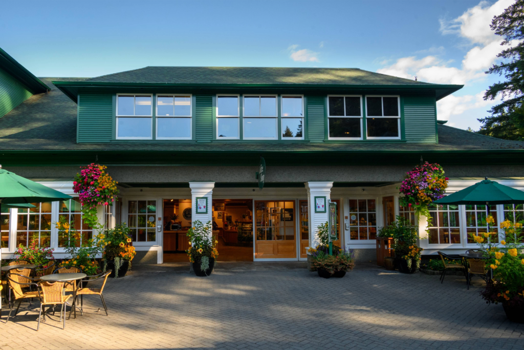 A green, two-story building with large windows and a sloped roof. The entrance features double doors flanked by potted plants and hanging flower baskets. Outdoor seating with tables and chairs is arranged in front, under green umbrellas