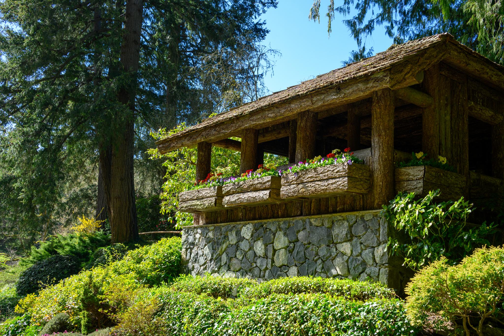 A rustic wooden gazebo with a thatched roof sits on a stone foundation, surrounded by lush greenery and colorful flower beds. The gazebo features a railing adorned with blooming flowers, while tall trees provide shade in the background