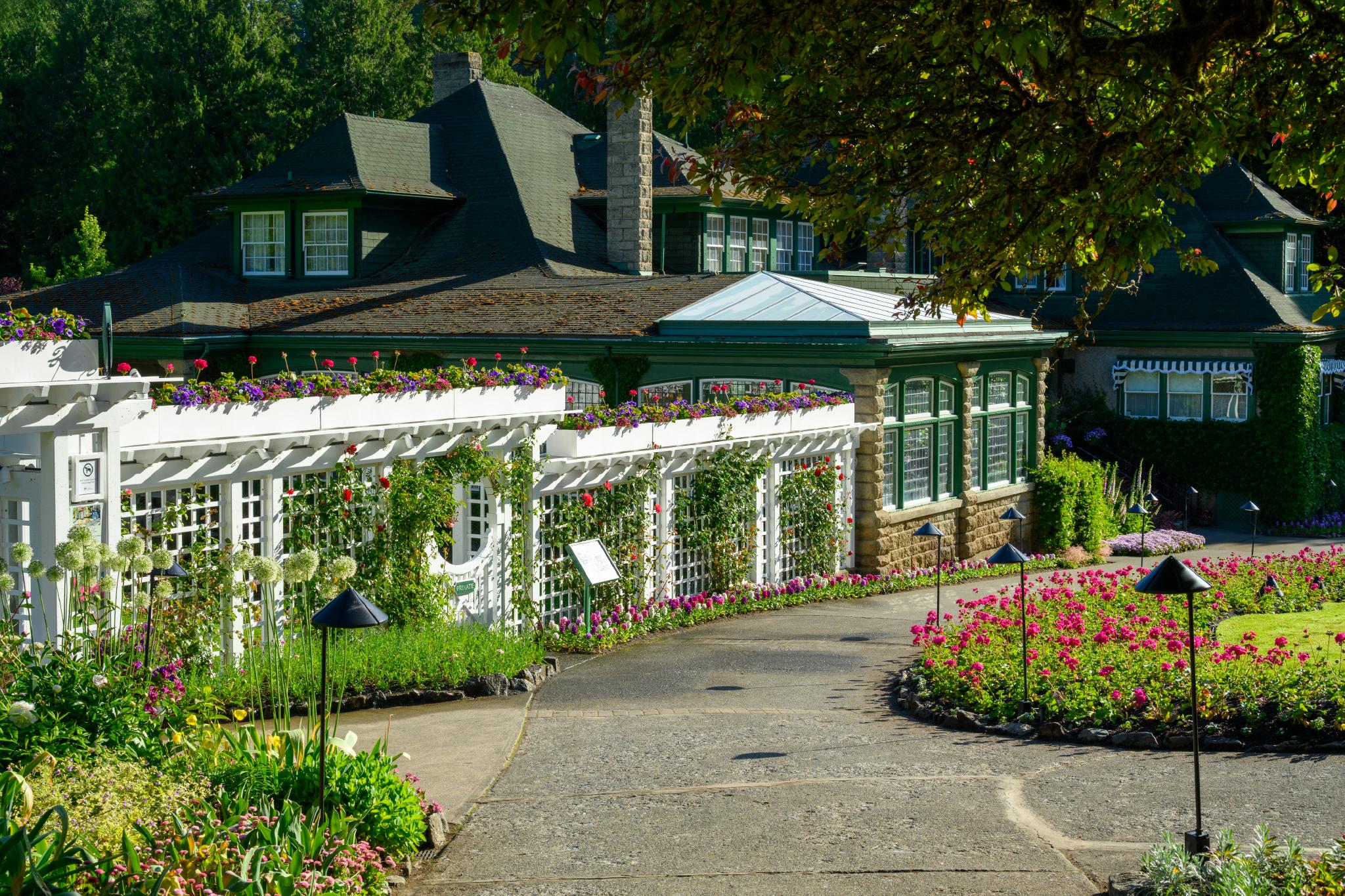 A landscaped garden featuring a pathway leading to a green, multi-windowed building. The garden includes colorful flower beds with various blooming plants, bordered by white trellises adorned with climbing vines and hanging flowers. The surrounding area has well-maintained grass and decorative lighting fixtures