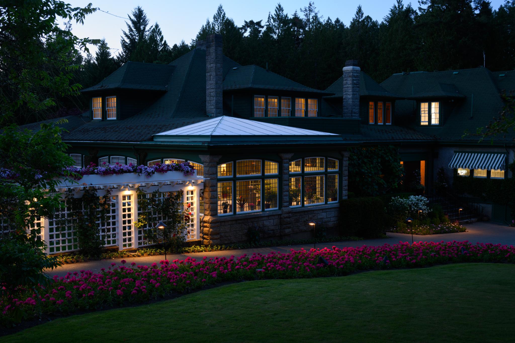 A large, two-story house with a dark green roof and multiple windows, illuminated by warm lights. The front features a garden with a neatly trimmed lawn and a flower bed filled with pink and purple blooms. Surrounding trees are visible in the background, indicating a wooded area