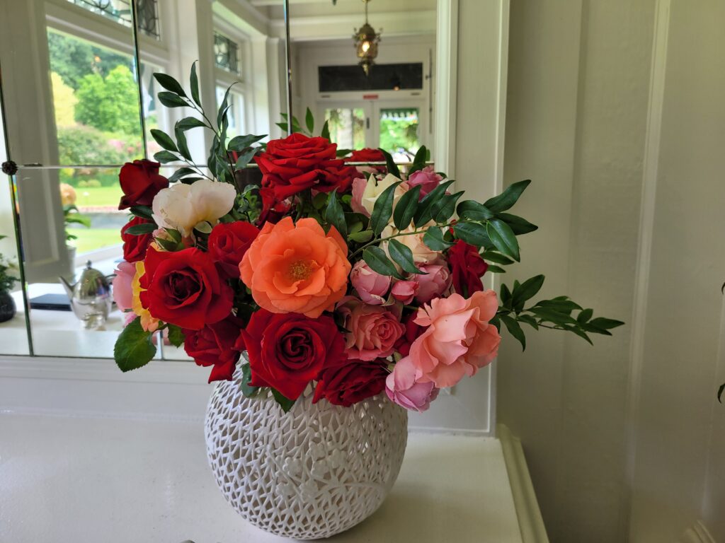 A bouquet of various roses, including red, pink, and orange blooms, arranged in a textured white vase. The roses display a range of sizes and shades, with lush green leaves interspersed among the flowers. The background features a bright, well-lit room with a mirror and greenery visible outside