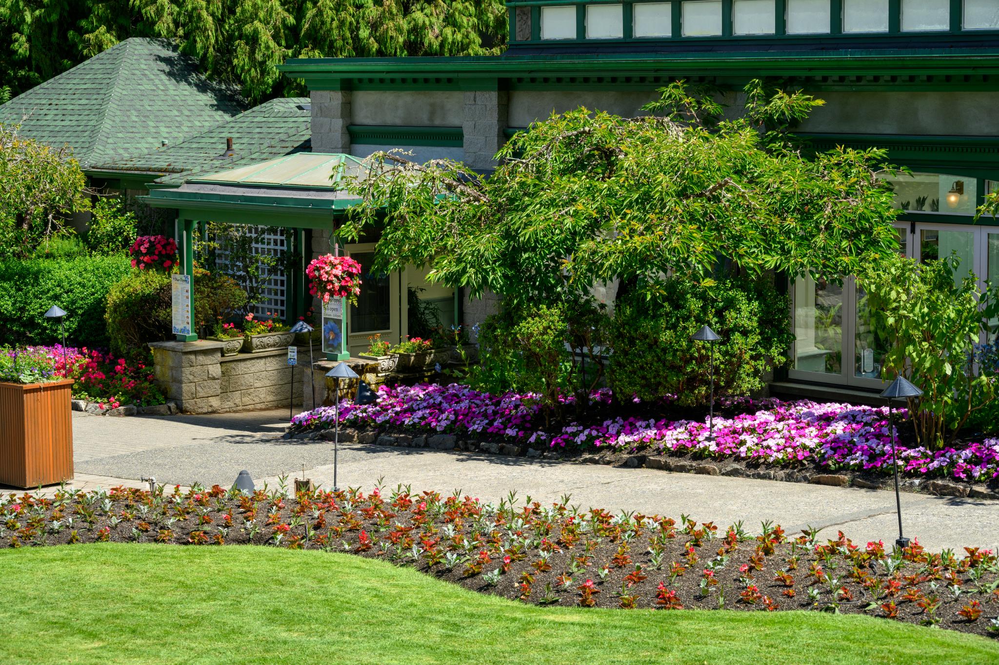 A landscaped garden featuring a variety of colorful flowers, including pink and white impatiens (genus Impatiens) in the foreground. The background shows a building with green trim and a porch adorned with hanging flower baskets. Lush greenery surrounds the area, creating a vibrant and well-maintained outdoor space