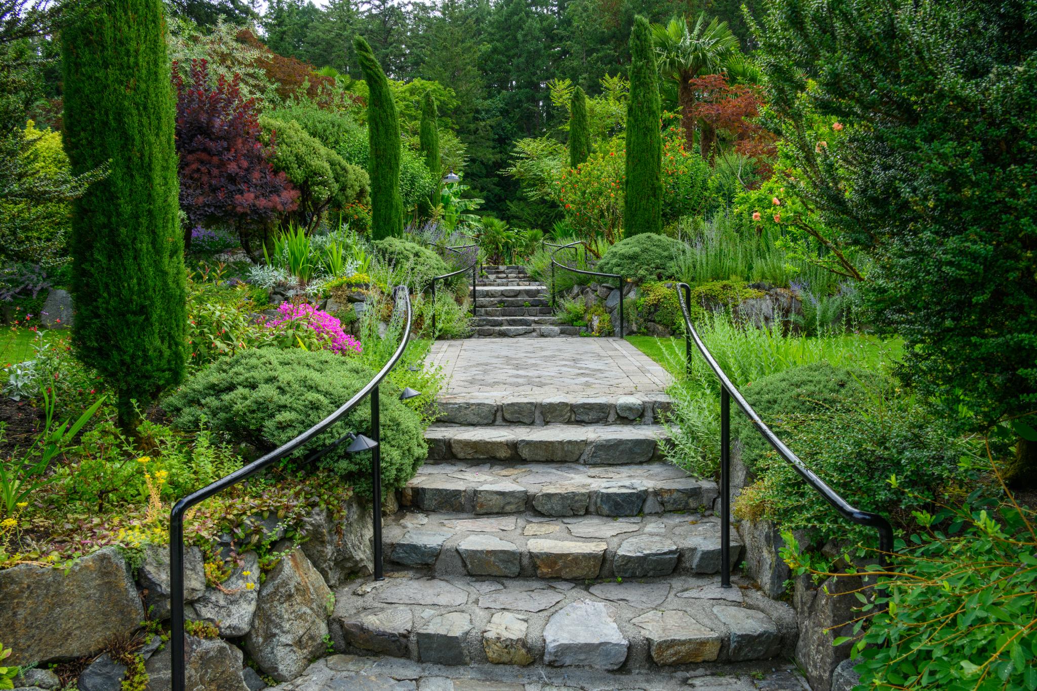 A stone pathway with metal railings leads through a lush garden filled with various plants, including coniferous trees and flowering shrubs. The garden features a mix of greenery, with vibrant colors from blooming flowers and ornamental plants, set against a backdrop of trees and foliage