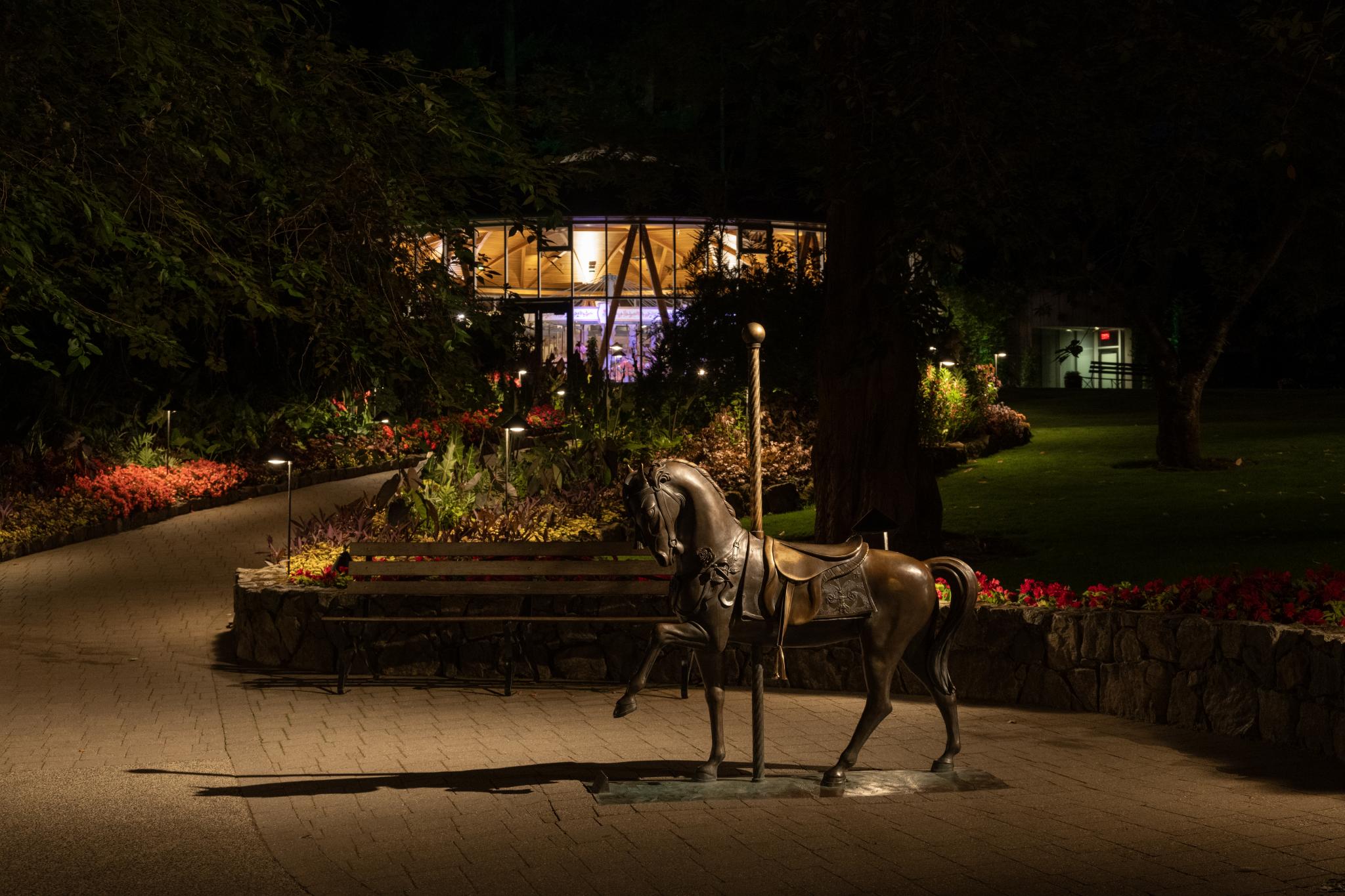 A bronze statue of a horse stands on a paved path, illuminated by soft lighting. In the background, a circular building with large windows is partially visible, surrounded by colorful flower beds and greenery. The scene is set at night, with the path leading towards the building