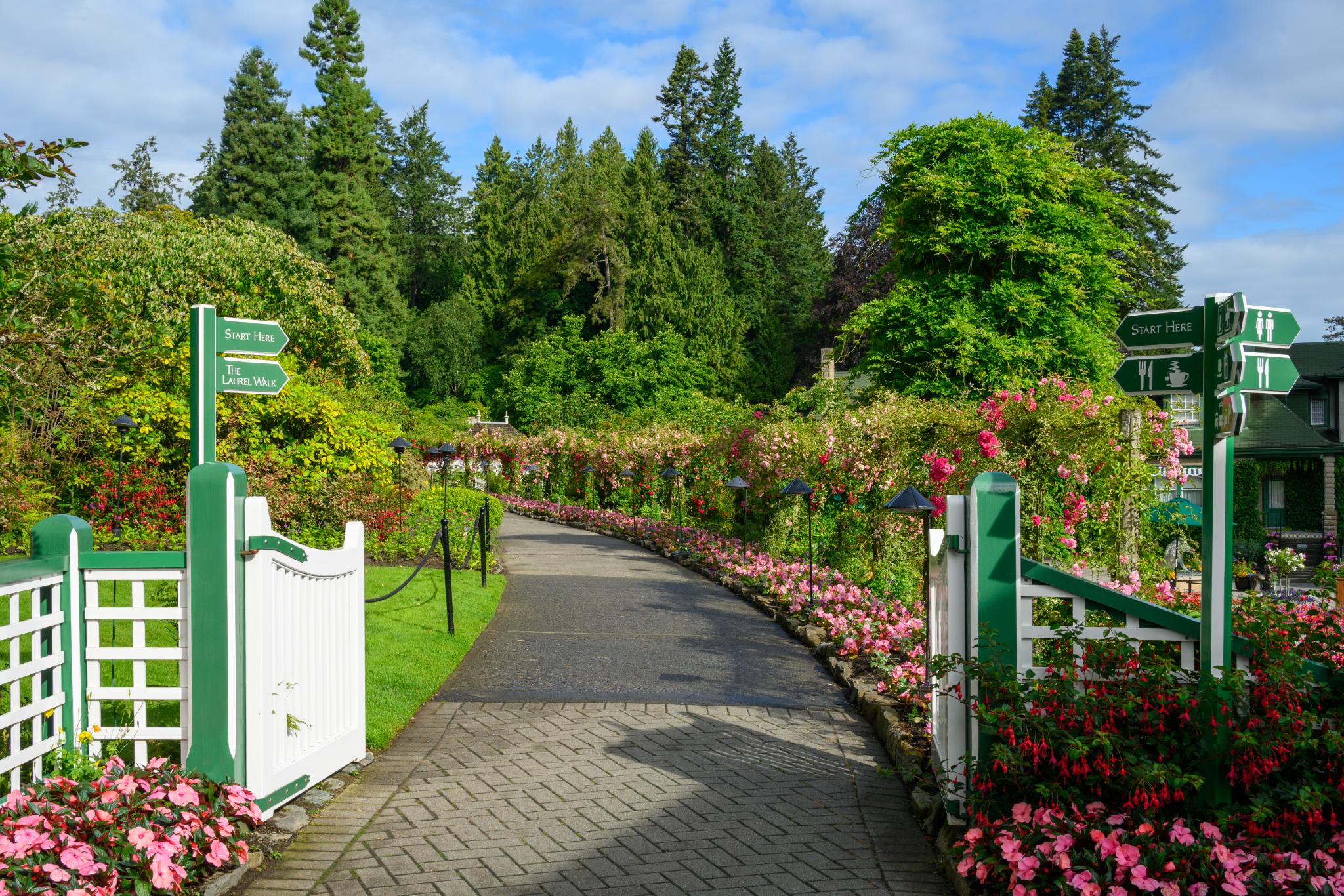 A winding pathway lined with colorful flowers and greenery, leading through a garden. The path is bordered by white fences and green signage indicating directions. Lush trees and shrubs are visible in the background, contributing to a serene outdoor setting