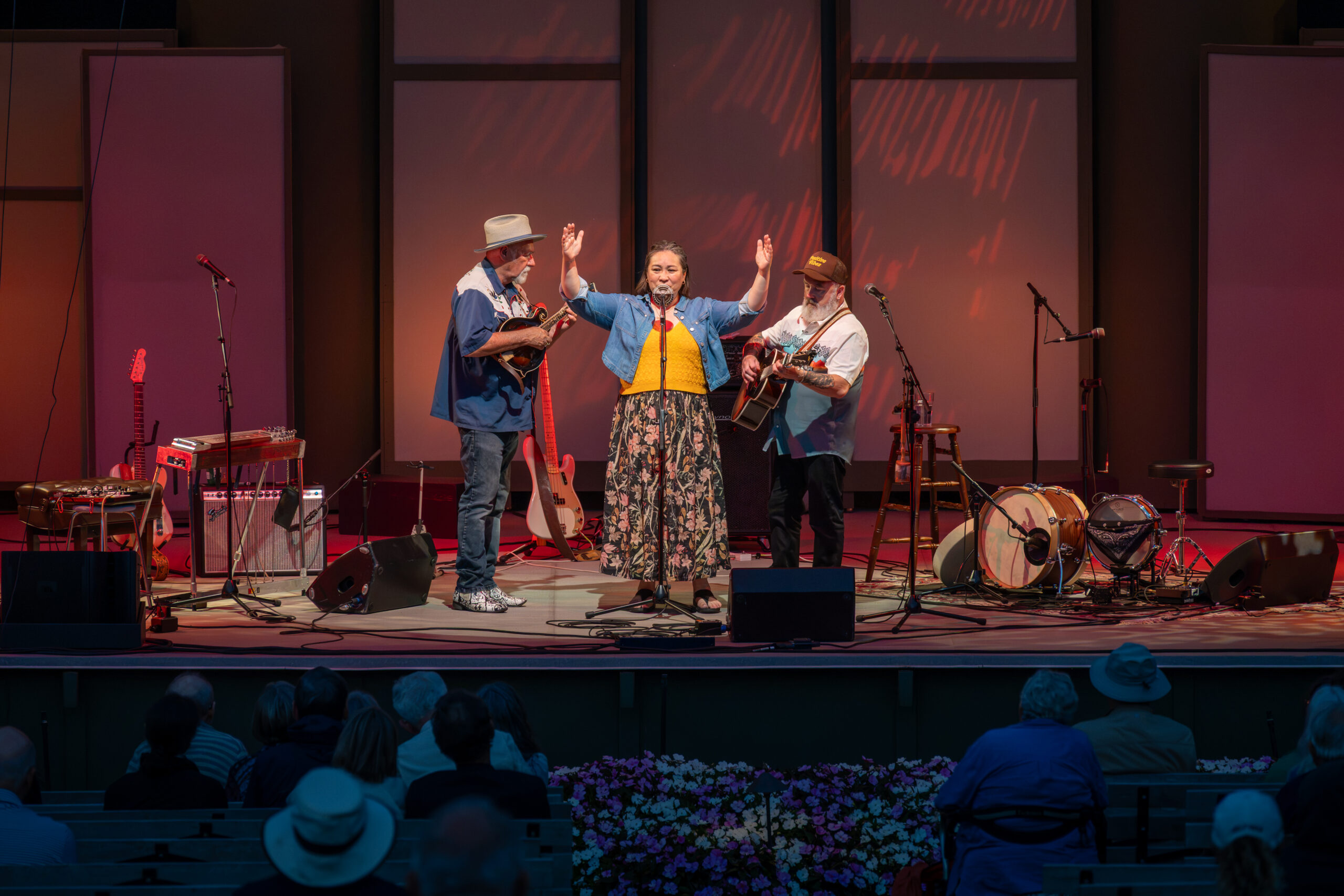 A woman in a colorful dress stands center stage with her arms raised, singing. She is flanked by two musicians: one playing a mandolin and the other holding a guitar. The stage is set with various musical instruments and microphones, and the audience is seated in front, partially visible. The lighting creates a warm atmosphere with red and orange hues