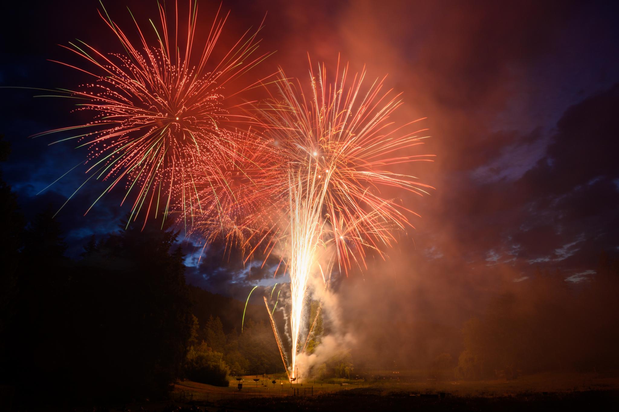 Brightly colored fireworks burst in the night sky, displaying vibrant reds and golds against a dark blue backdrop. The fireworks are set off from a ground location, with trails of light and smoke visible. The scene captures the excitement and illumination of a festive celebration
