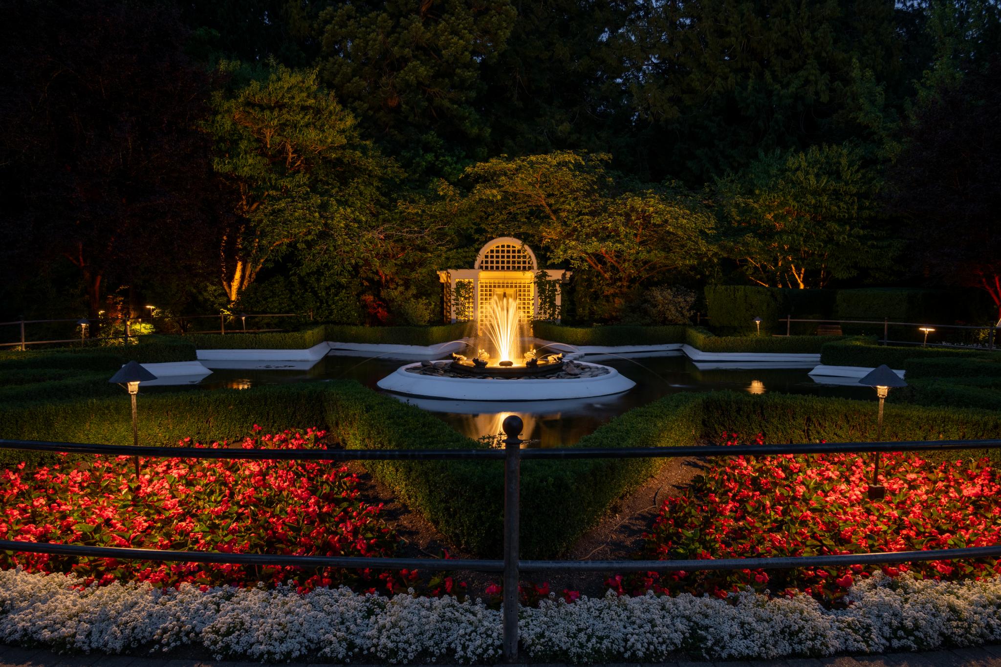 A formal garden at night featuring a central fountain illuminated by warm light. Surrounding the fountain are neatly arranged flower beds with red and white blooms, bordered by low hedges. Soft lighting highlights the garden's pathways and structures, creating a serene atmosphere