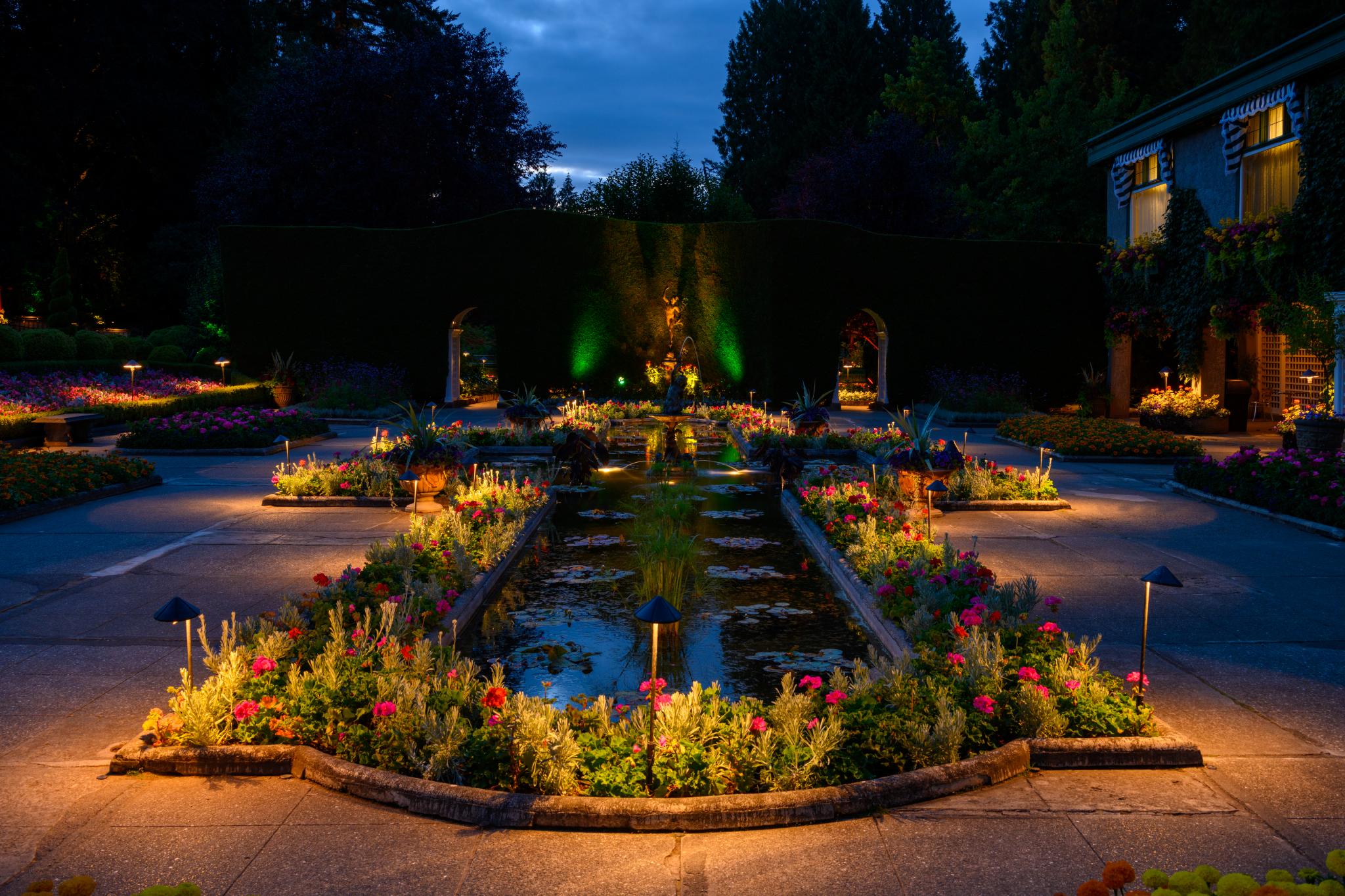 A landscaped garden at dusk, featuring a central pond surrounded by flower beds. The beds contain a variety of flowering plants, with visible colors including pink, yellow, and purple. Soft lighting illuminates the pathways and plants, creating a serene atmosphere. Green hedges form a backdrop, and decorative structures are present on either side of the garden