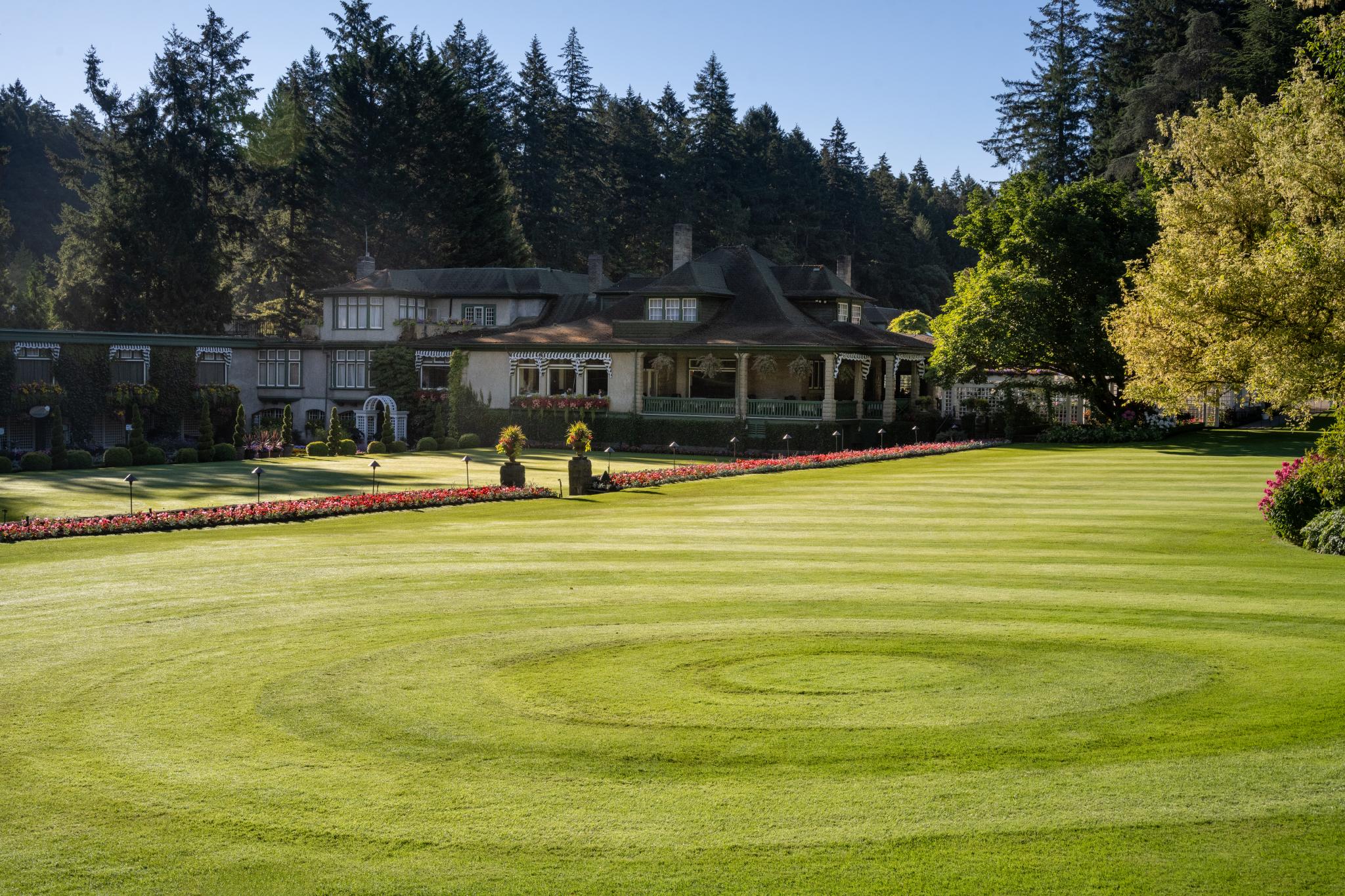 A well-manicured lawn features a spiral pattern cut into the grass, leading towards a large, elegant house surrounded by trees. Colorful flower beds line the edges of the lawn, adding visual interest to the landscape
