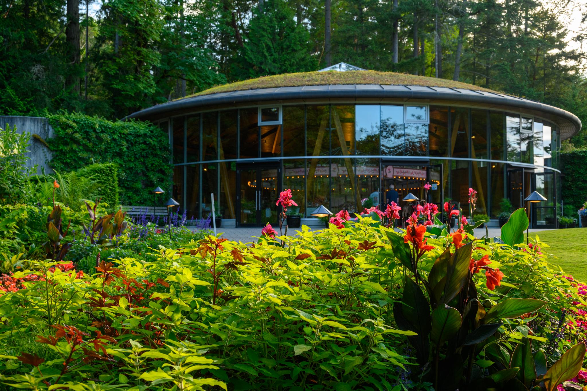 A round, modern building with large glass windows is surrounded by vibrant green foliage and colorful flowers. In the foreground, there are clusters of red and orange flowers, possibly from the genus Canna, alongside lush green leaves. The building features a green roof and is set against a backdrop of trees
