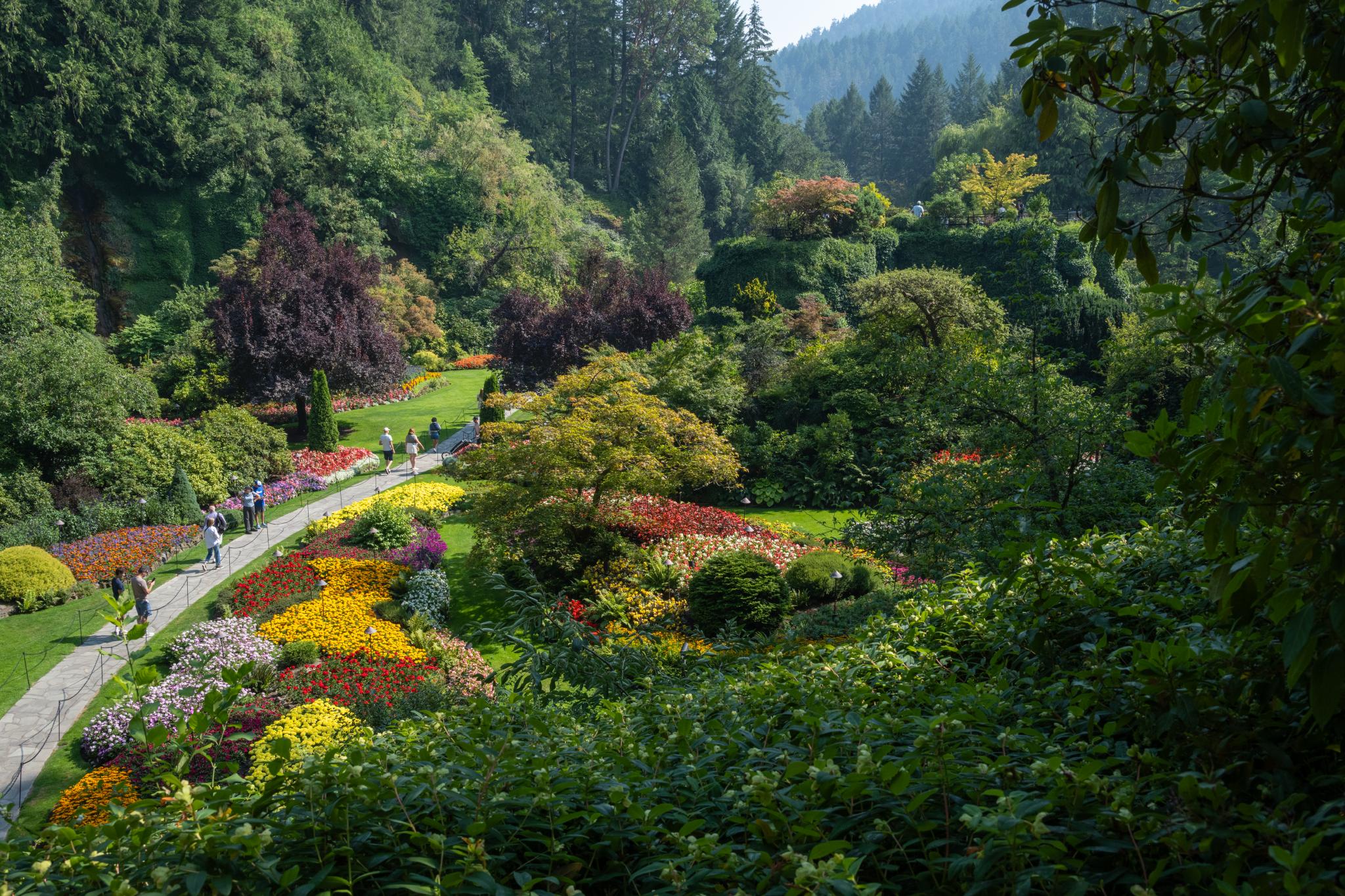 A landscaped garden featuring a variety of flowering plants and shrubs, with vibrant colors including reds, yellows, and purples. The scene includes winding pathways and visitors walking through the garden, surrounded by lush greenery and trees in the background