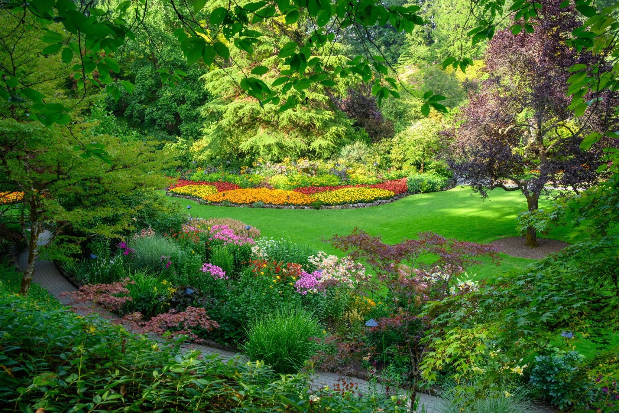 A landscaped garden featuring a variety of flowering plants and shrubs. The scene includes vibrant flower beds arranged in circular patterns with colors ranging from yellow to red, surrounded by lush green grass and trees. The garden is framed by greenery, creating a serene and colorful outdoor space