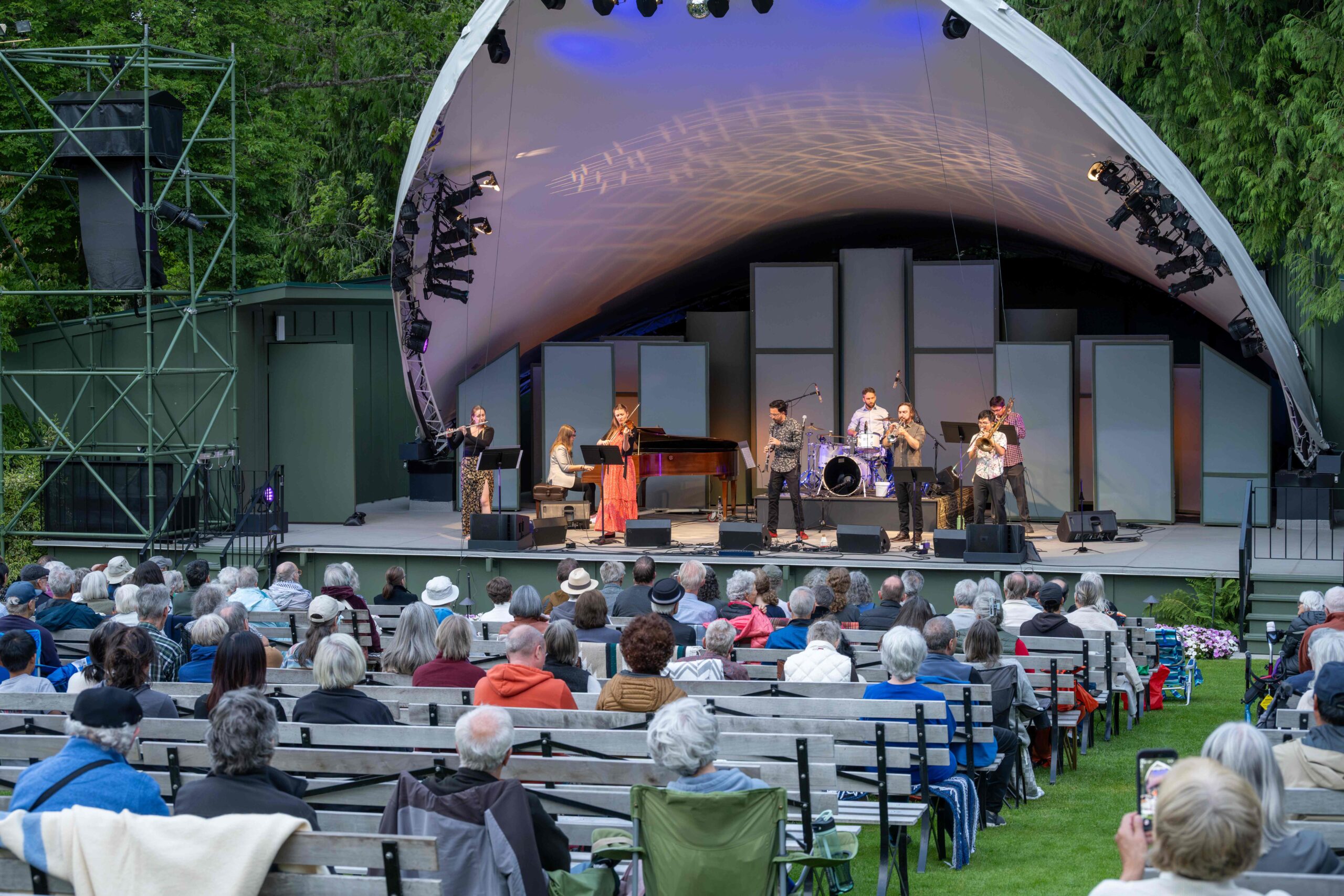 A live music performance is taking place on an outdoor stage with a large, curved canopy. The performers include a band with various instruments, and a seated audience is visible in front, watching the show. The crowd is composed of diverse individuals, some wearing hats and enjoying the event. The stage is set against a backdrop of greenery