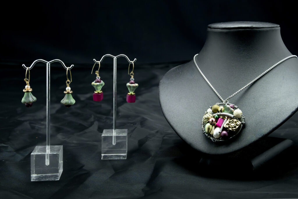 Deux paires de boucles d'oreilles et un collier présentés sur un fond noir. Les boucles d'oreilles, suspendues à des supports transparents, arborent des motifs floraux agrémentés d'éléments verts et violets. Le collier, posé sur un buste noir, met en valeur un pendentif rond orné de diverses perles et de motifs floraux dans des tons de vert, de blanc et de violet.