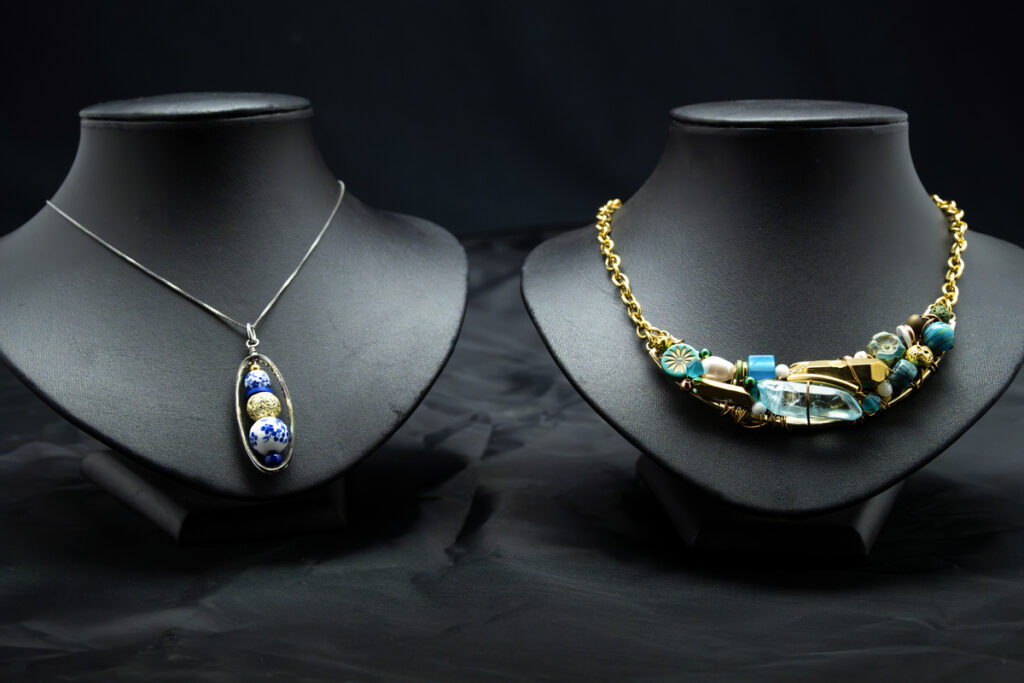 Deux présentoirs à bijoux posés sur un tissu noir. À gauche, un collier en argent orné d'un pendentif ovale au motif bleu et or. À droite, un collier à grosse chaîne en or agrémenté de perles turquoise et transparentes, ainsi que d'éléments décoratifs.