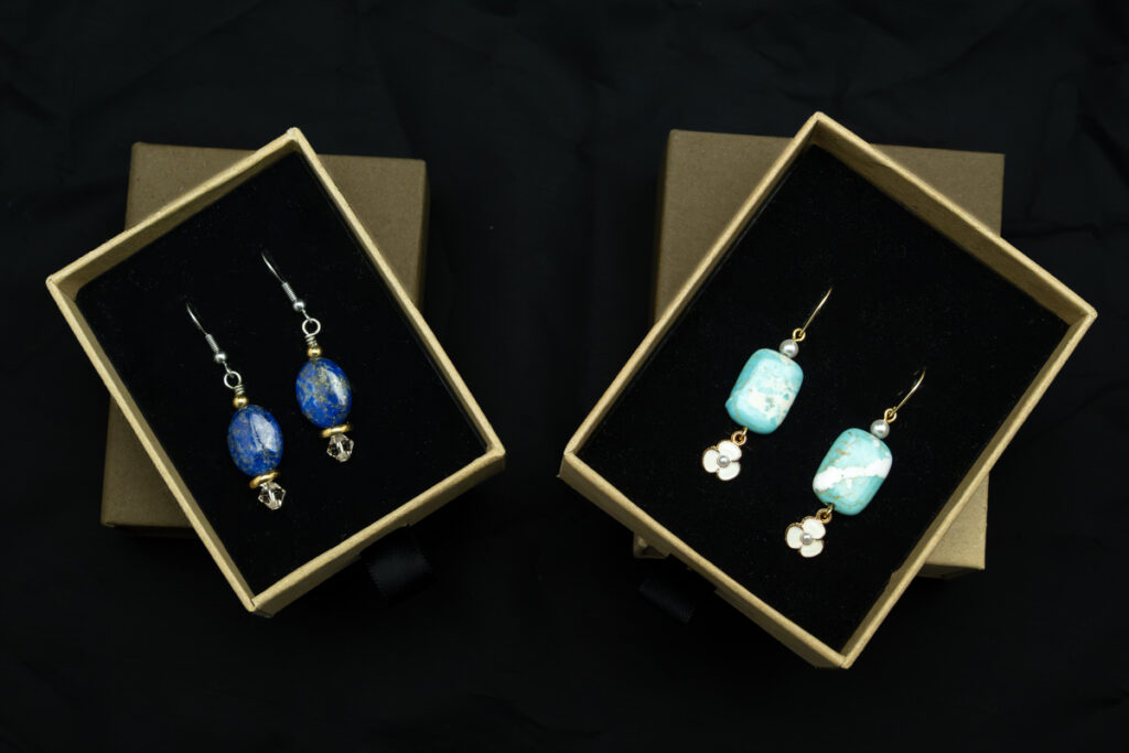 Deux paires de boucles d'oreilles présentées dans des boîtes à bijoux ouvertes. La paire de gauche est composée de perles bleues à la surface lisse, tandis que celle de droite comporte des perles bleu clair à l'aspect marbré ; toutes deux sont suspendues à des crochets dorés. Les boîtes sont doublées de tissu noir et posées sur un fond sombre.