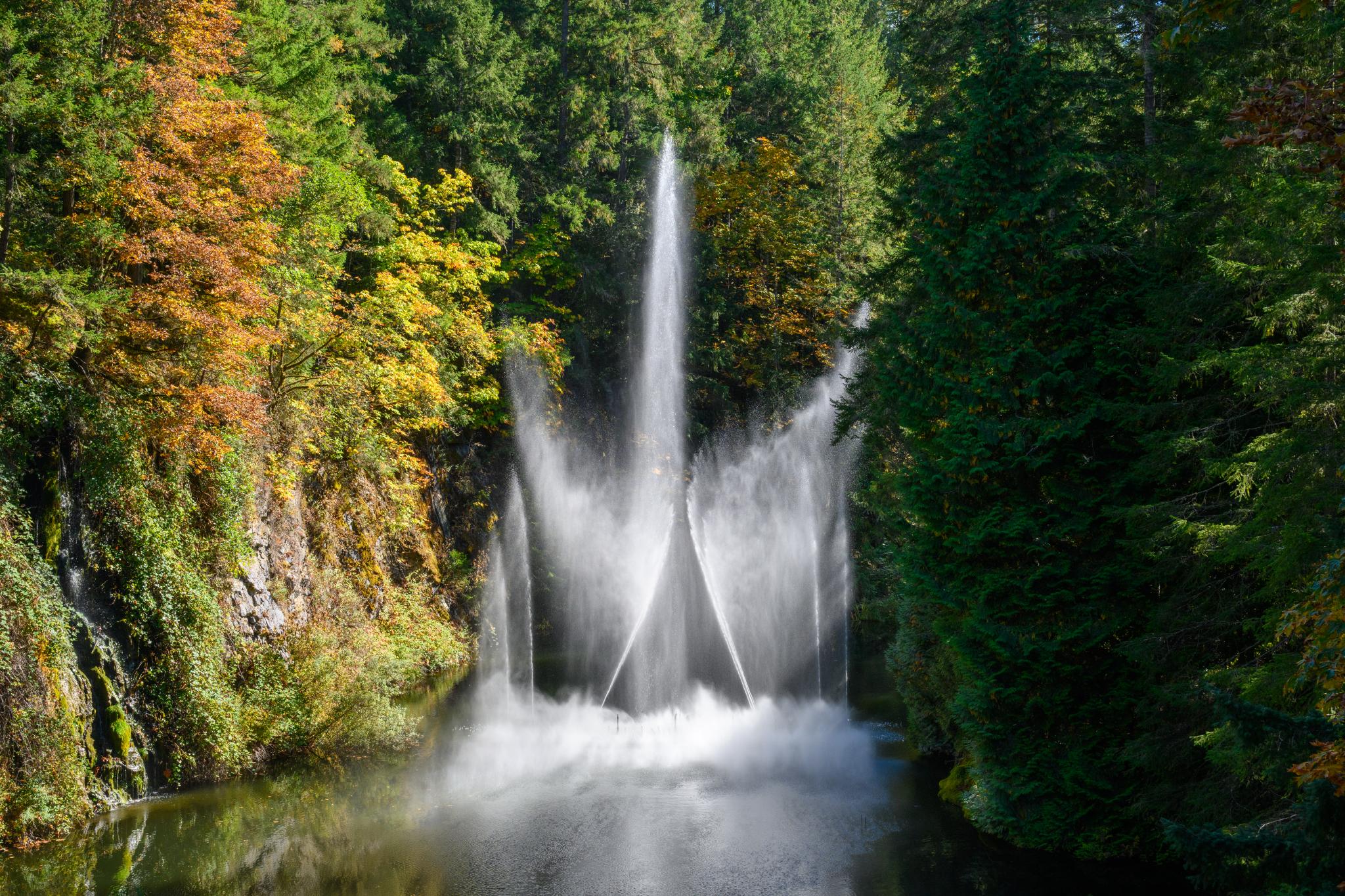 A waterfall cascades down rocky cliffs into a serene pool, surrounded by lush green coniferous trees and autumn foliage in shades of orange and yellow. The water creates a mist as it falls, enhancing the tranquil atmosphere of the scene