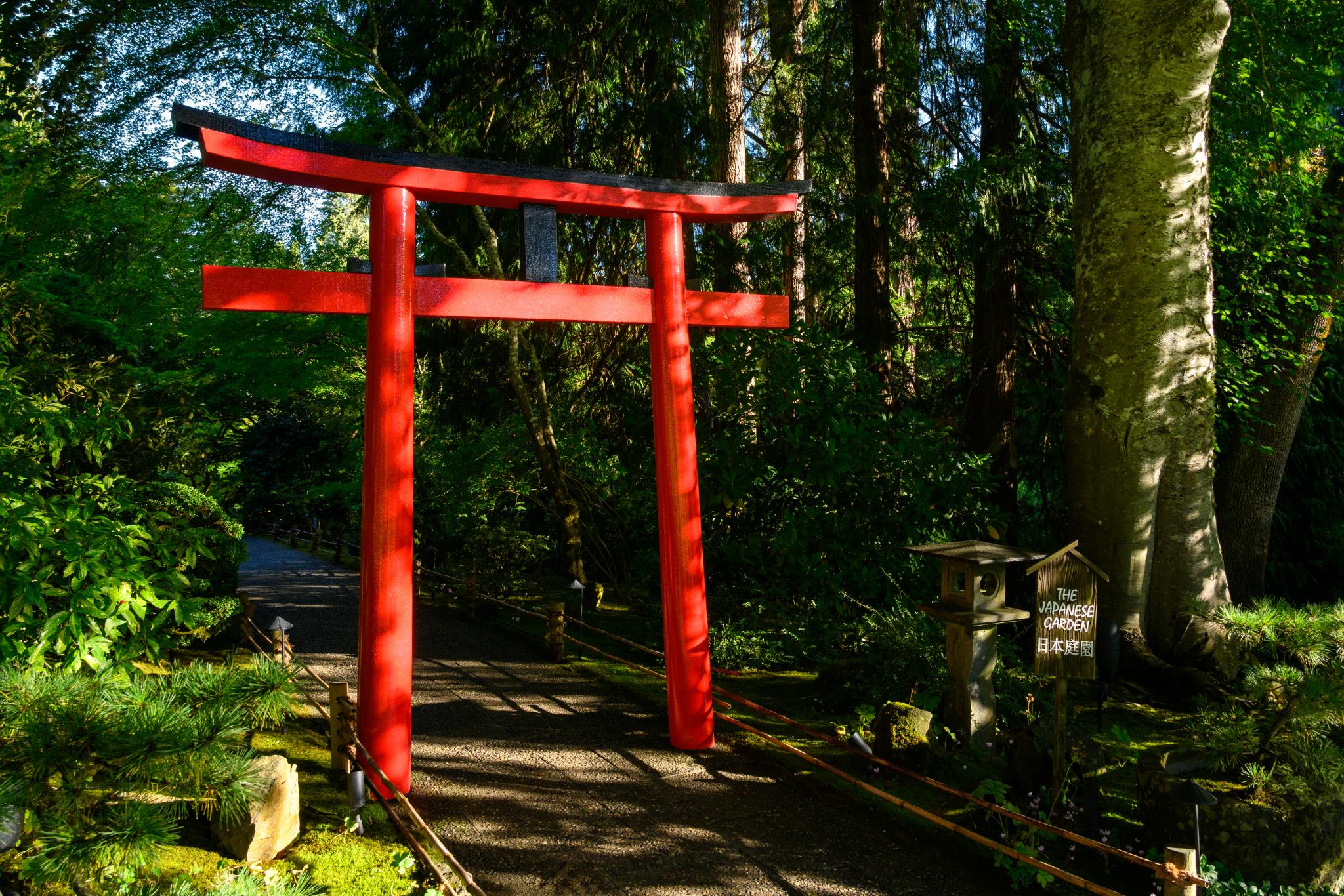 A large red torii gate stands at the entrance to a pathway surrounded by lush green foliage and trees. The gate is traditional in style, with two vertical posts and a horizontal beam at the top. Nearby, a stone lantern and a wooden sign are visible, indicating the path leads to a garden or serene area