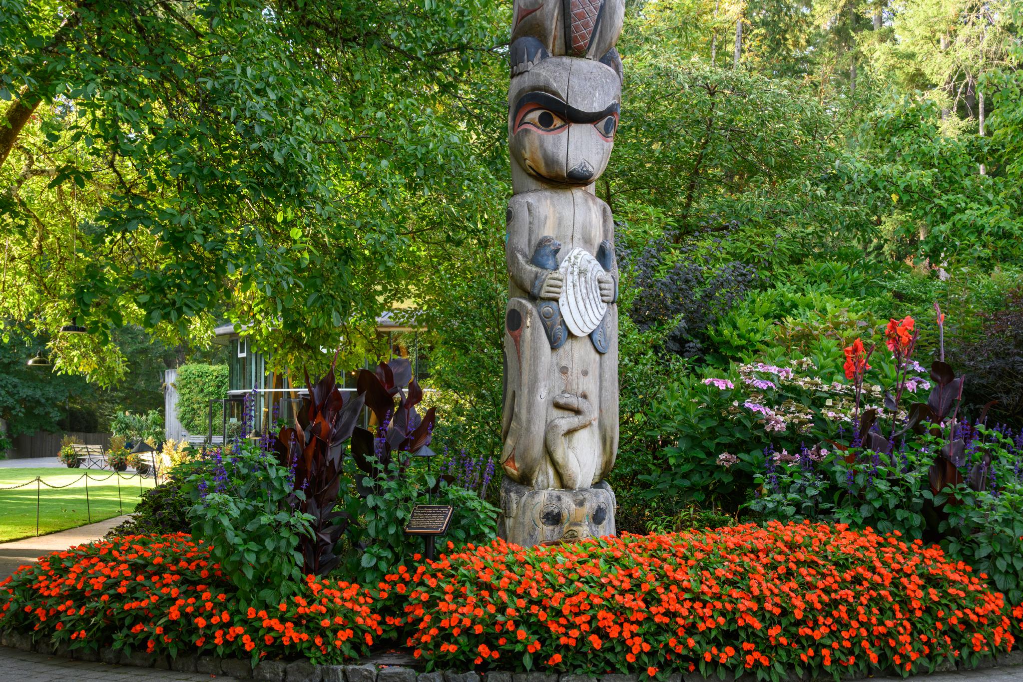 A wooden totem pole featuring a stylized figure with a prominent face and holding a shell, surrounded by a vibrant flower bed of orange flowers and various green plants. The background includes lush trees and shrubs, creating a natural setting